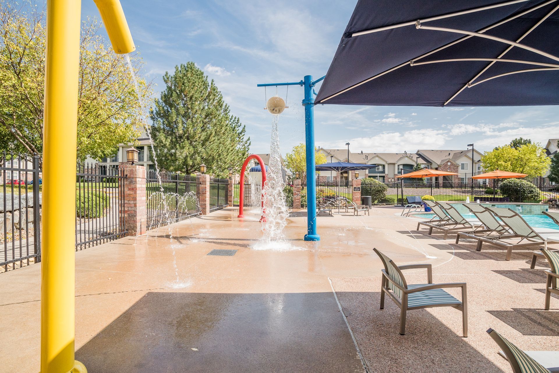 A splash pad area with water features, chairs, and an umbrella on a sunny day.