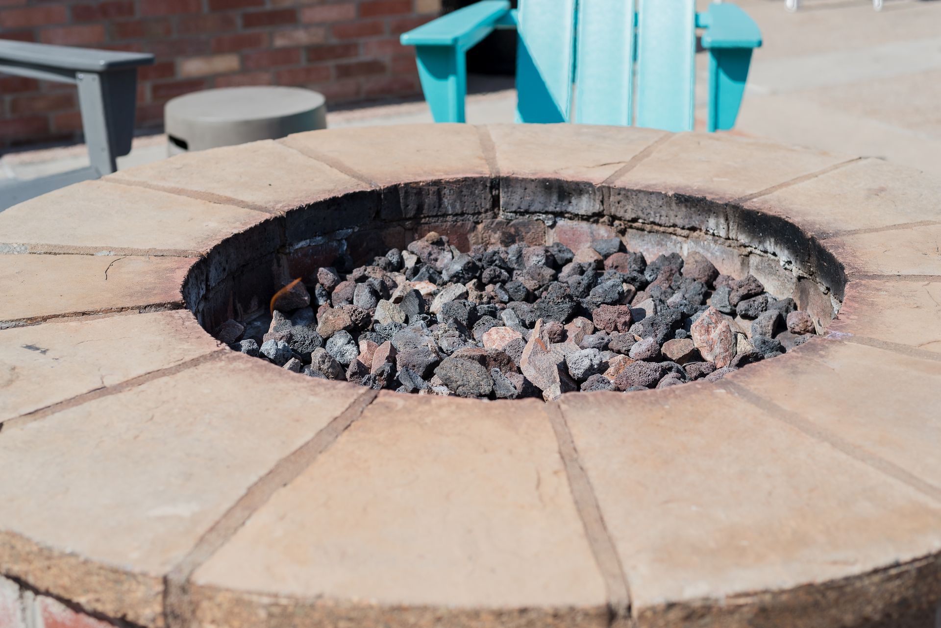 Fire pit with lava rocks, surrounded by light brick. Blue chair in background.
