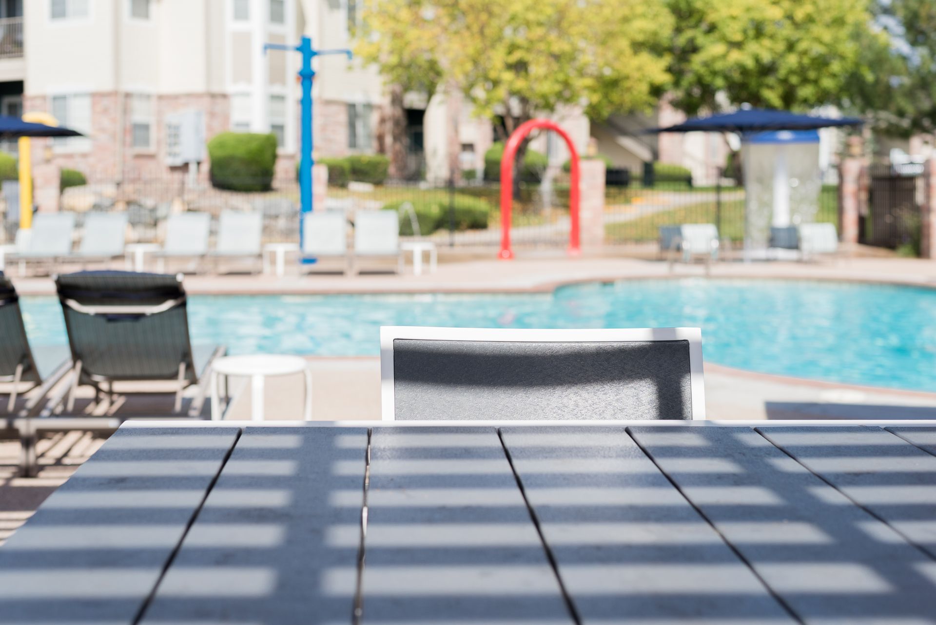 Poolside scene: gray table in foreground, lounge chairs, pool, water feature, apartment buildings in the background.