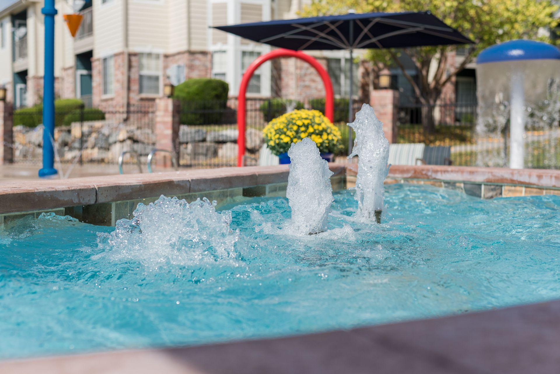 Fountains spray water in a blue pool. A splash pad with a blue and red arch and flowers in the background.