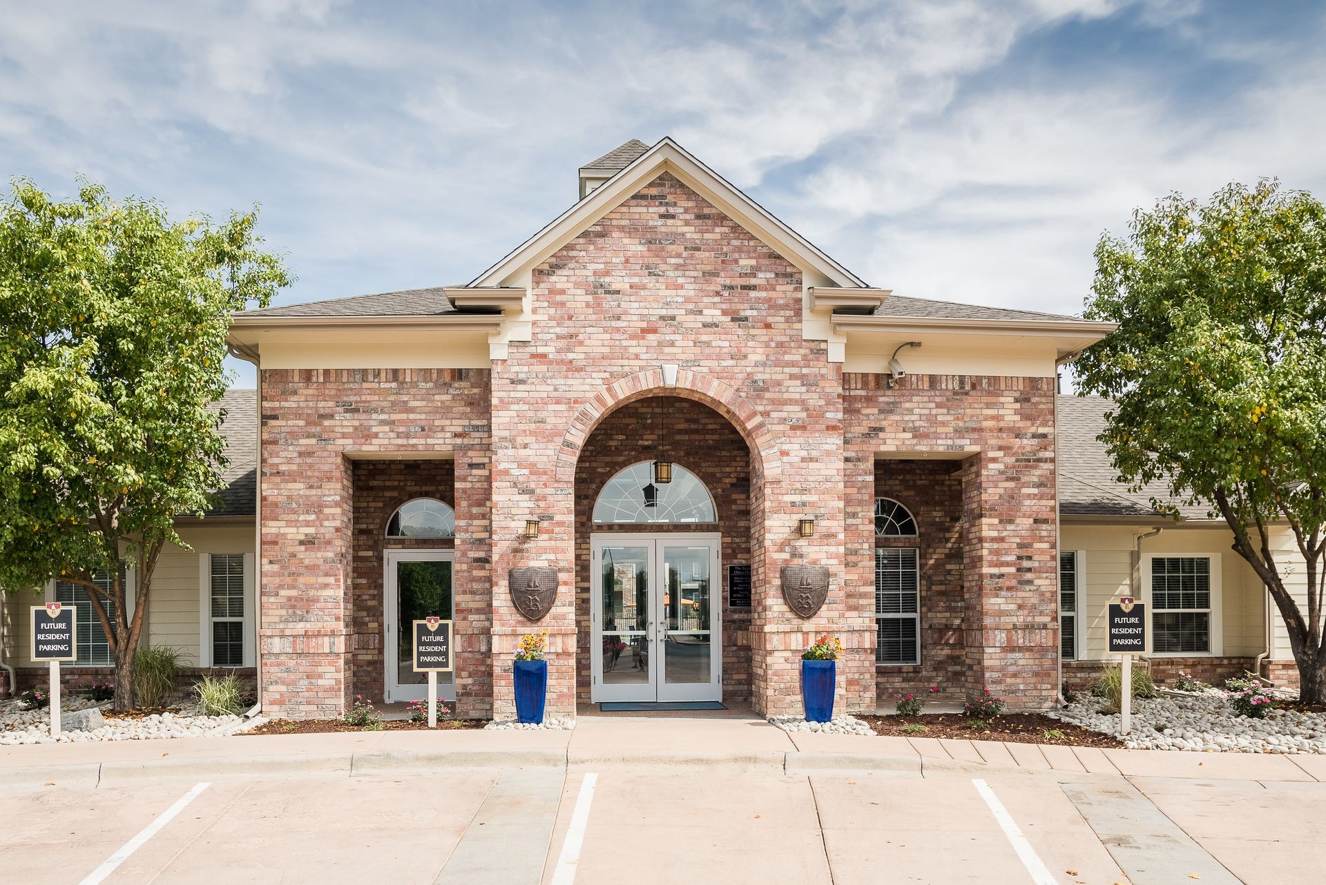 Brick building with arched entrance and double doors; blue planters, trees, and signs are present.