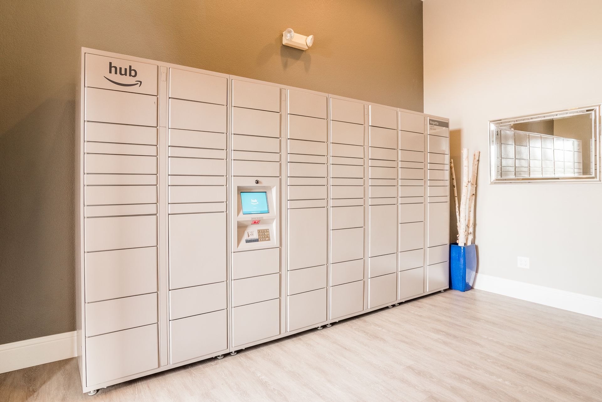 Silver Amazon Hub lockers in a hallway with a touchscreen, near a mirror and floor.