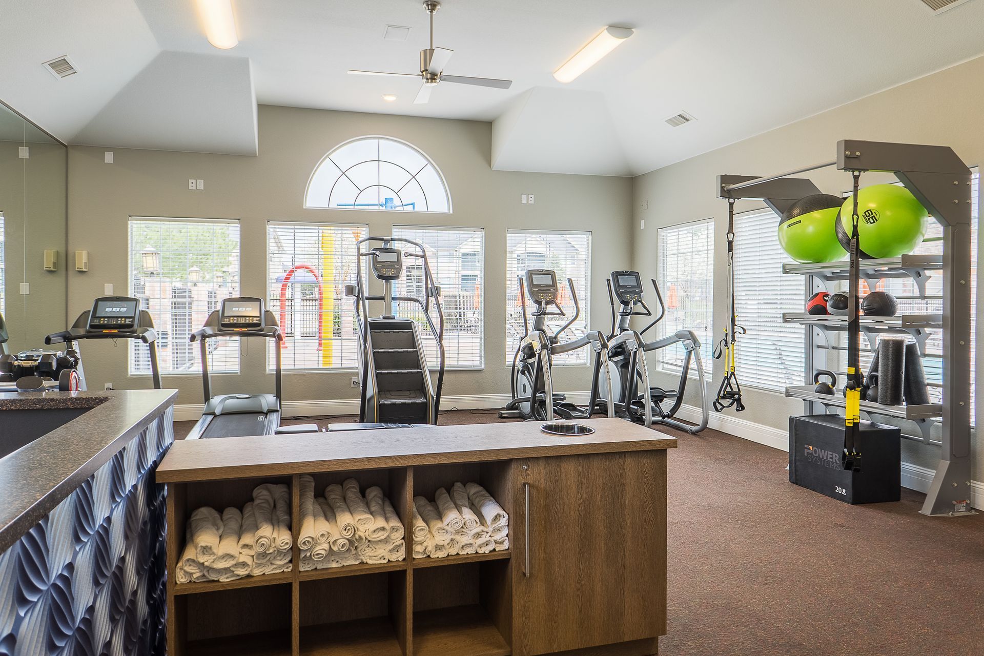 Gym interior with cardio machines, weight rack, and towels. Bright room with windows.