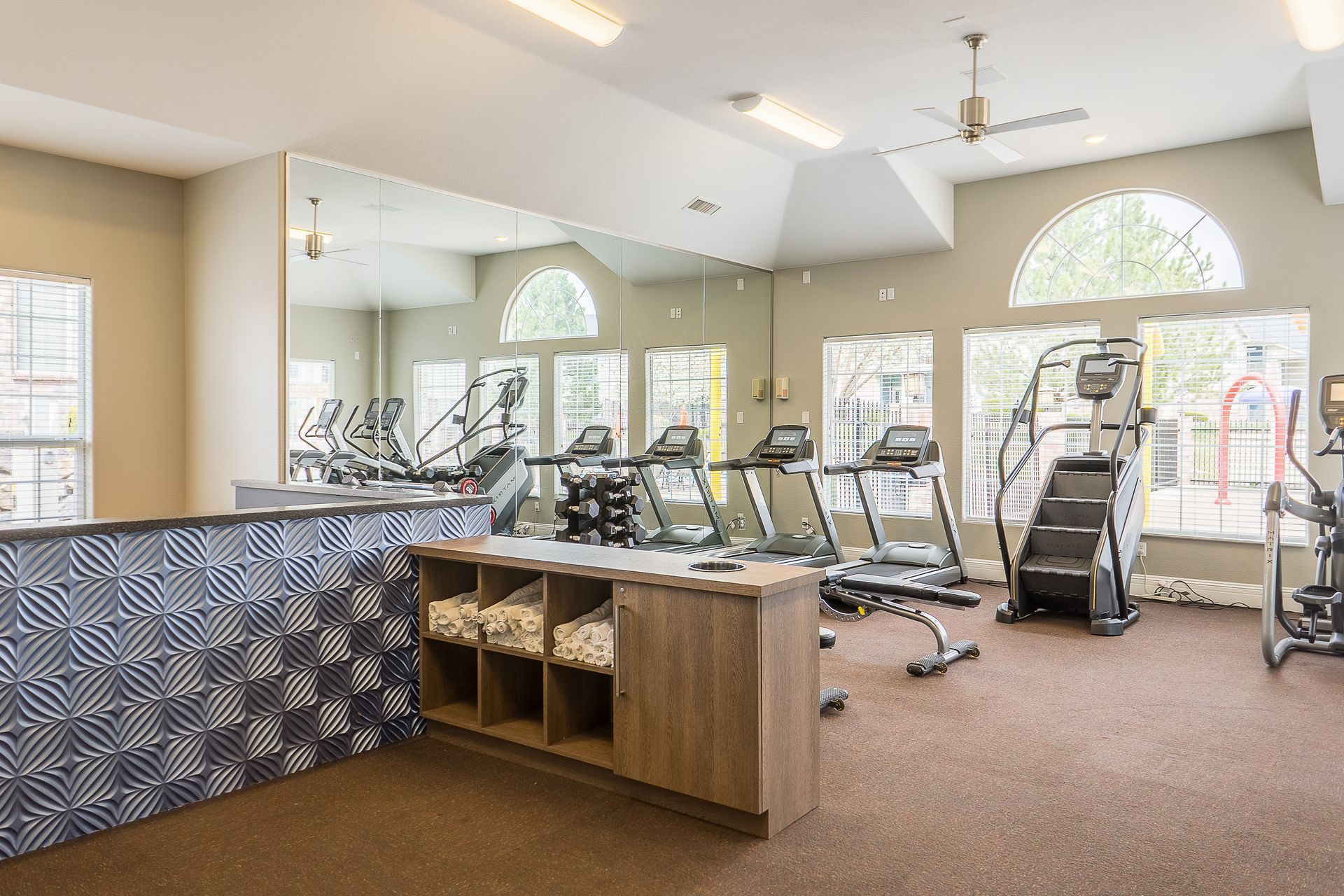 Gym with exercise equipment, towels, and large mirror. Beige walls, brown floor.