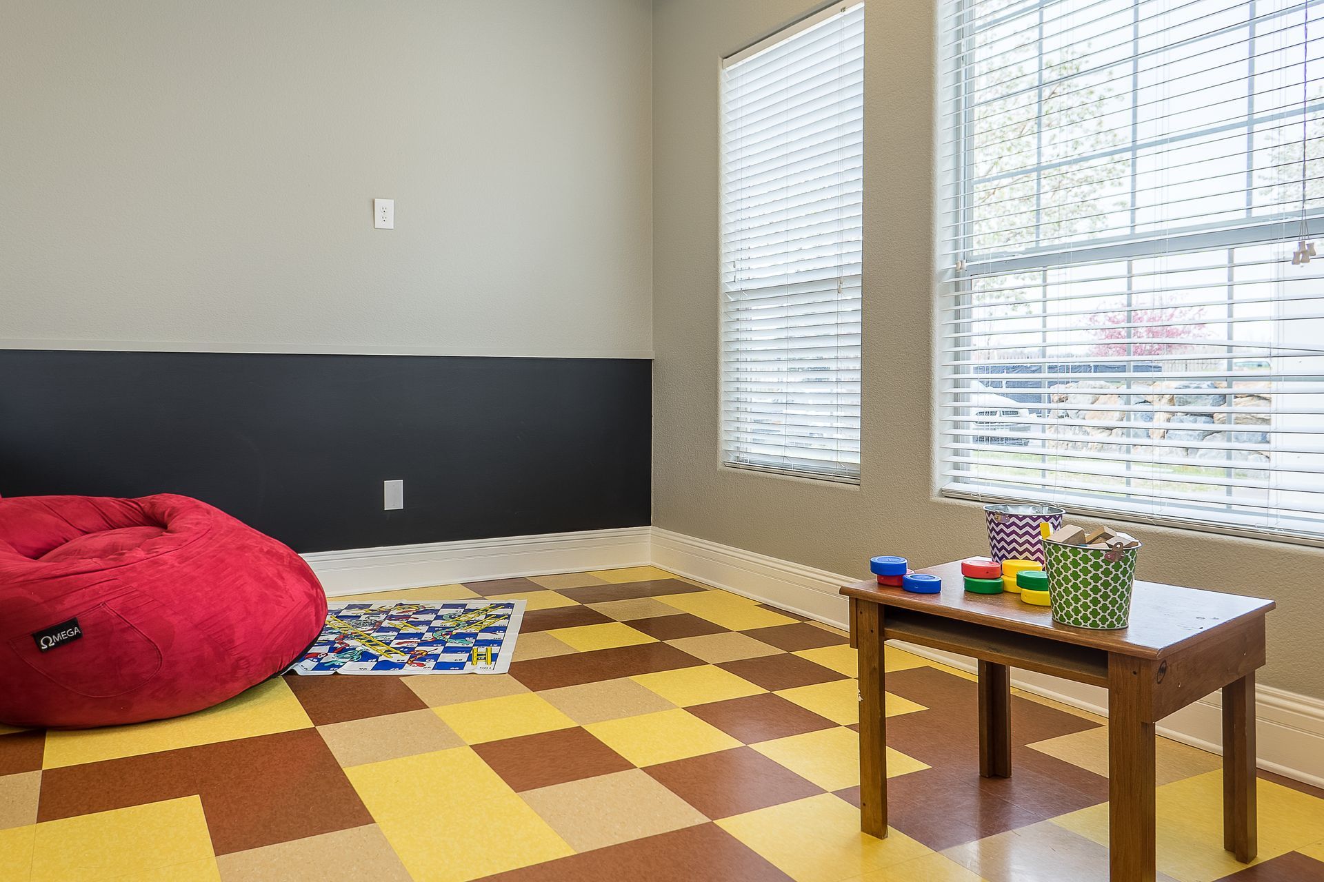 Children's play area with red beanbag chair, checkerboard floor, small table with toys, and windows.