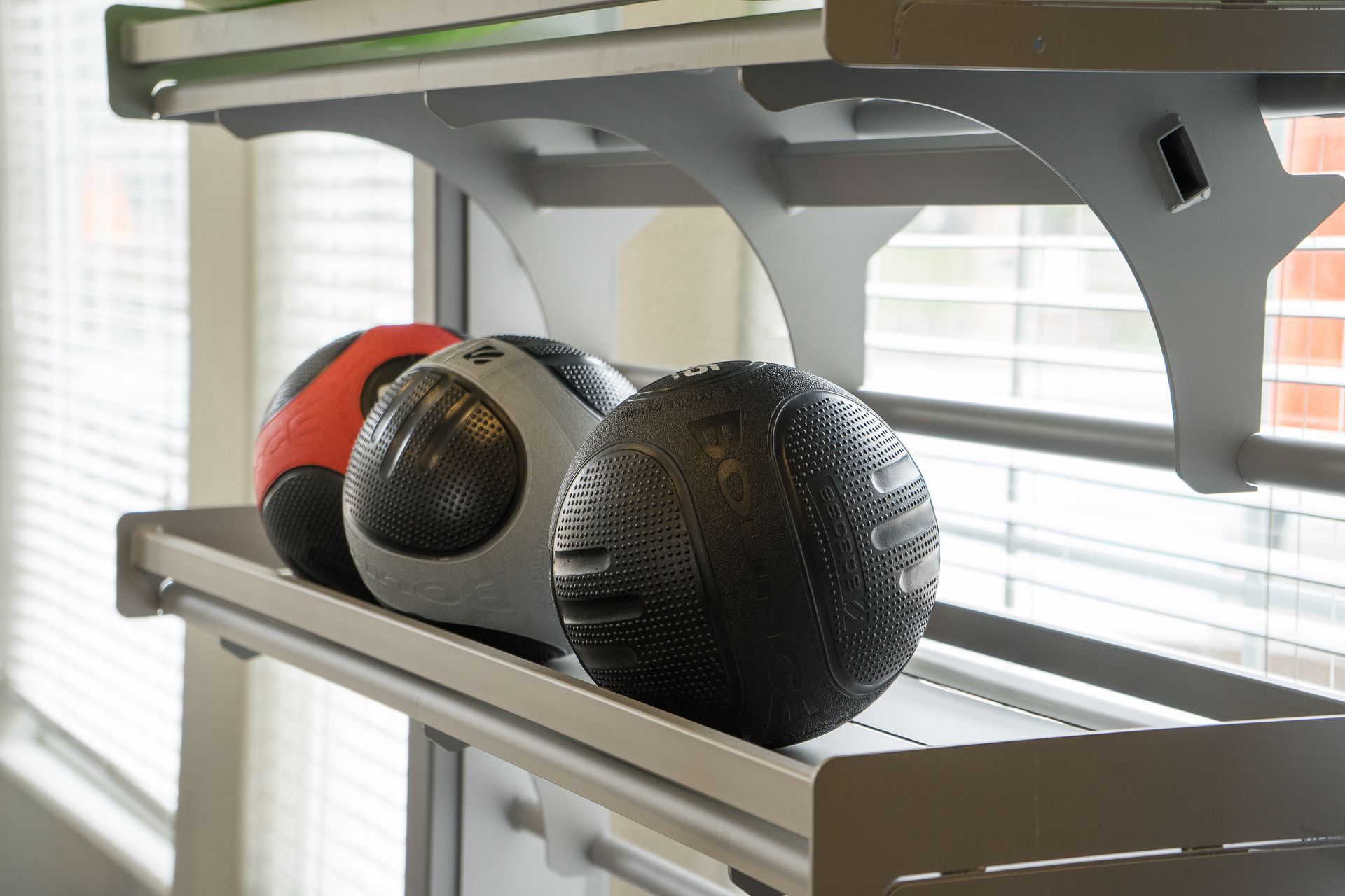 Three medicine balls, red, gray, and black, on a gym equipment shelf in front of a window.
