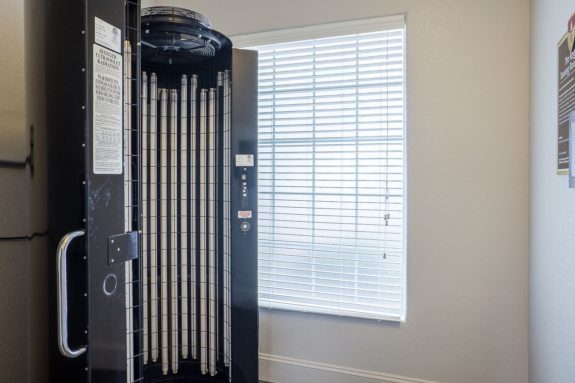 A tanning bed with open door, black, with long vertical lights. Window with blinds in background.