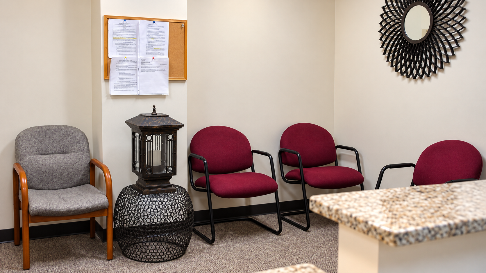 Waiting room with gray and burgundy chairs, a decorative lantern, bulletin board, and wall mirror.