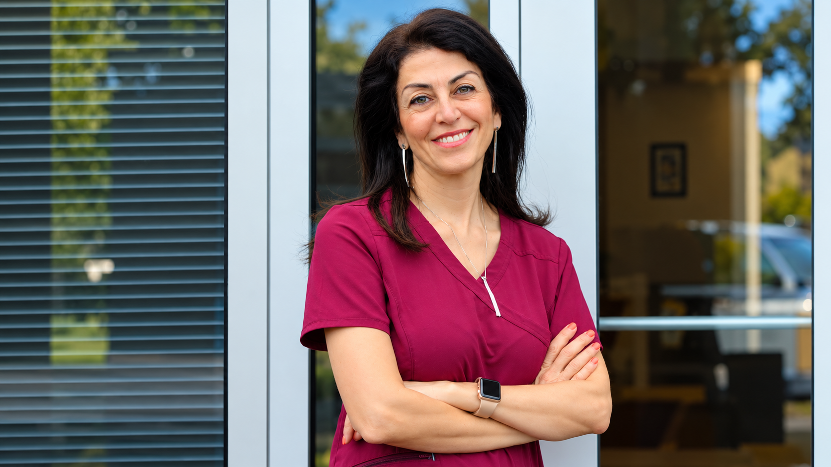 Woman in a magenta shirt stands smiling with arms crossed outside a building.