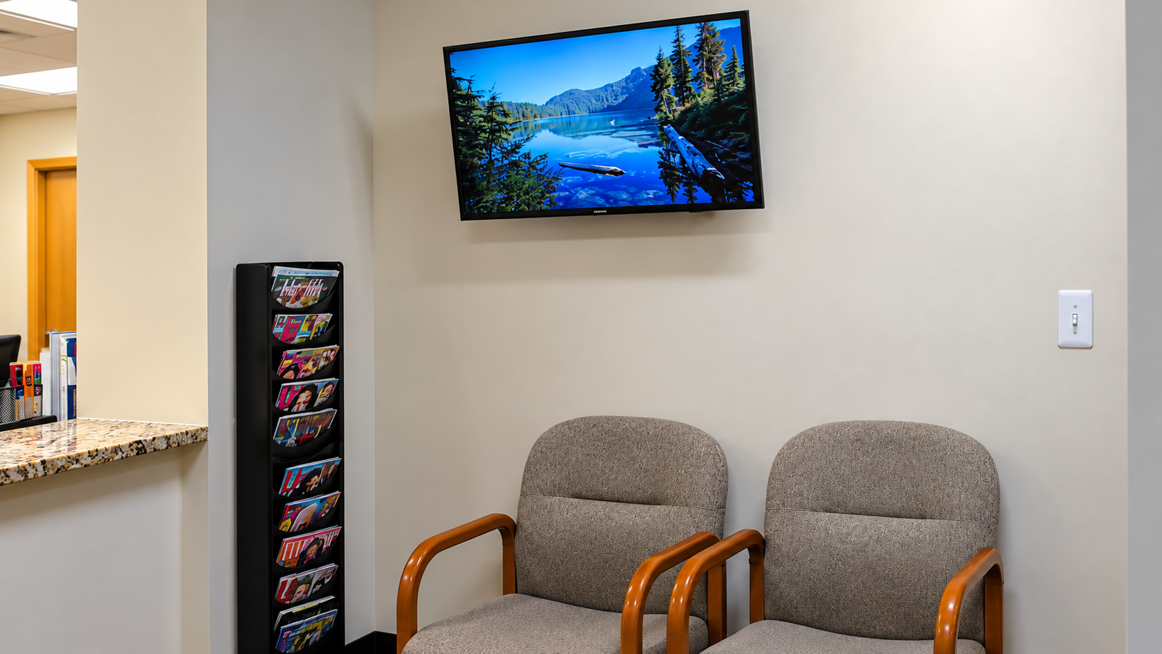 Waiting area with two chairs, wall-mounted TV, and brochure rack beside a reception counter.