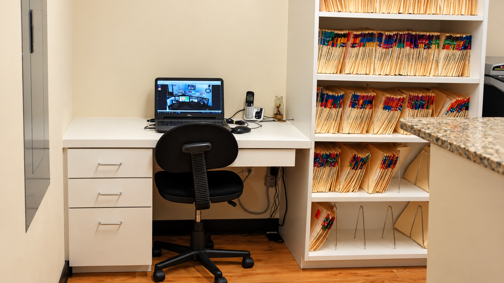 Small home office nook with white desk, black chair, laptop, and shelves of packaged snacks.