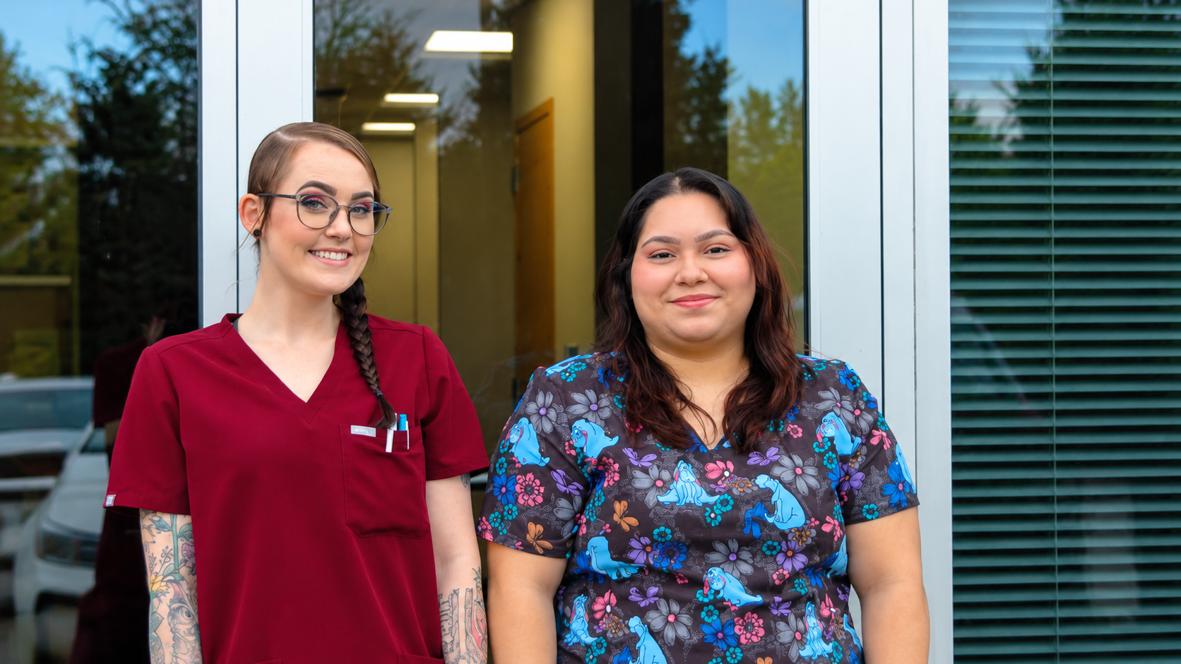 Two women standing outside a glass-door entrance, one in maroon scrubs and one in a floral top, smiling.