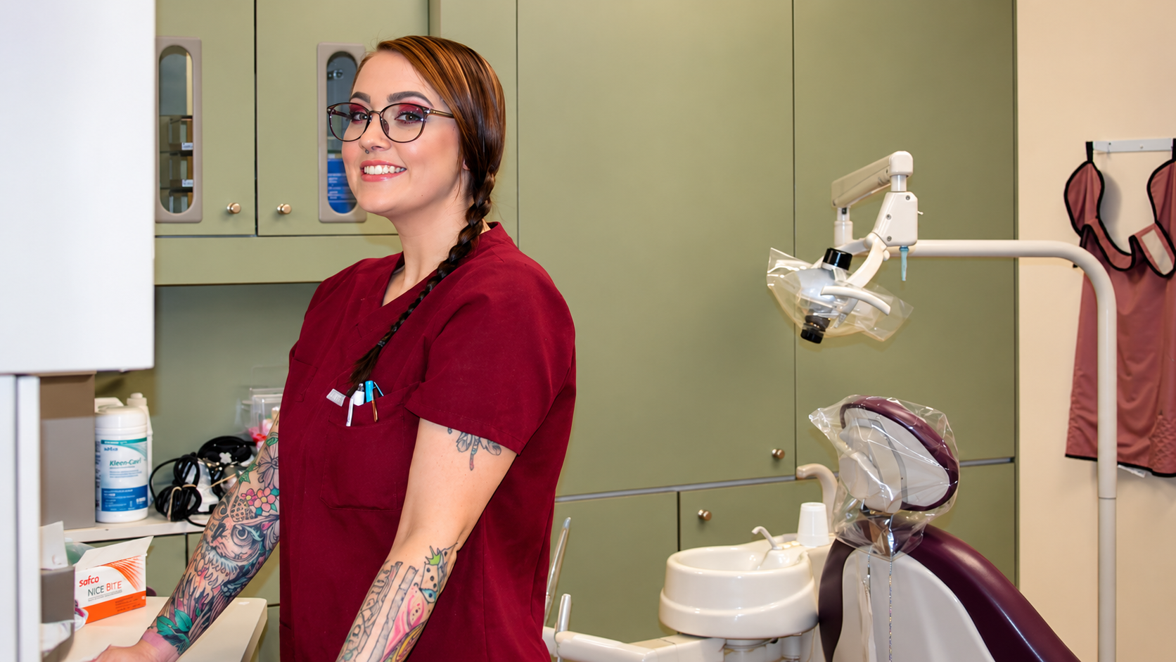 Dental hygienist in maroon scrubs smiling in a dental exam room beside equipment