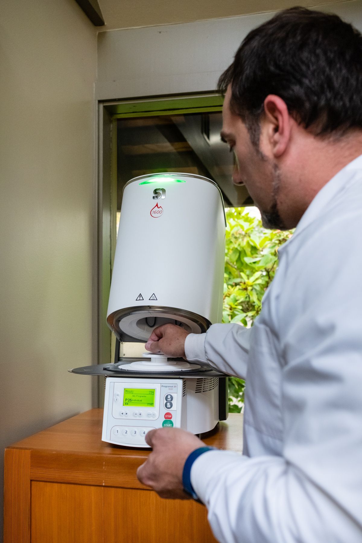 Man in lab coat operating a white furnace on a wooden surface, opening the lid.