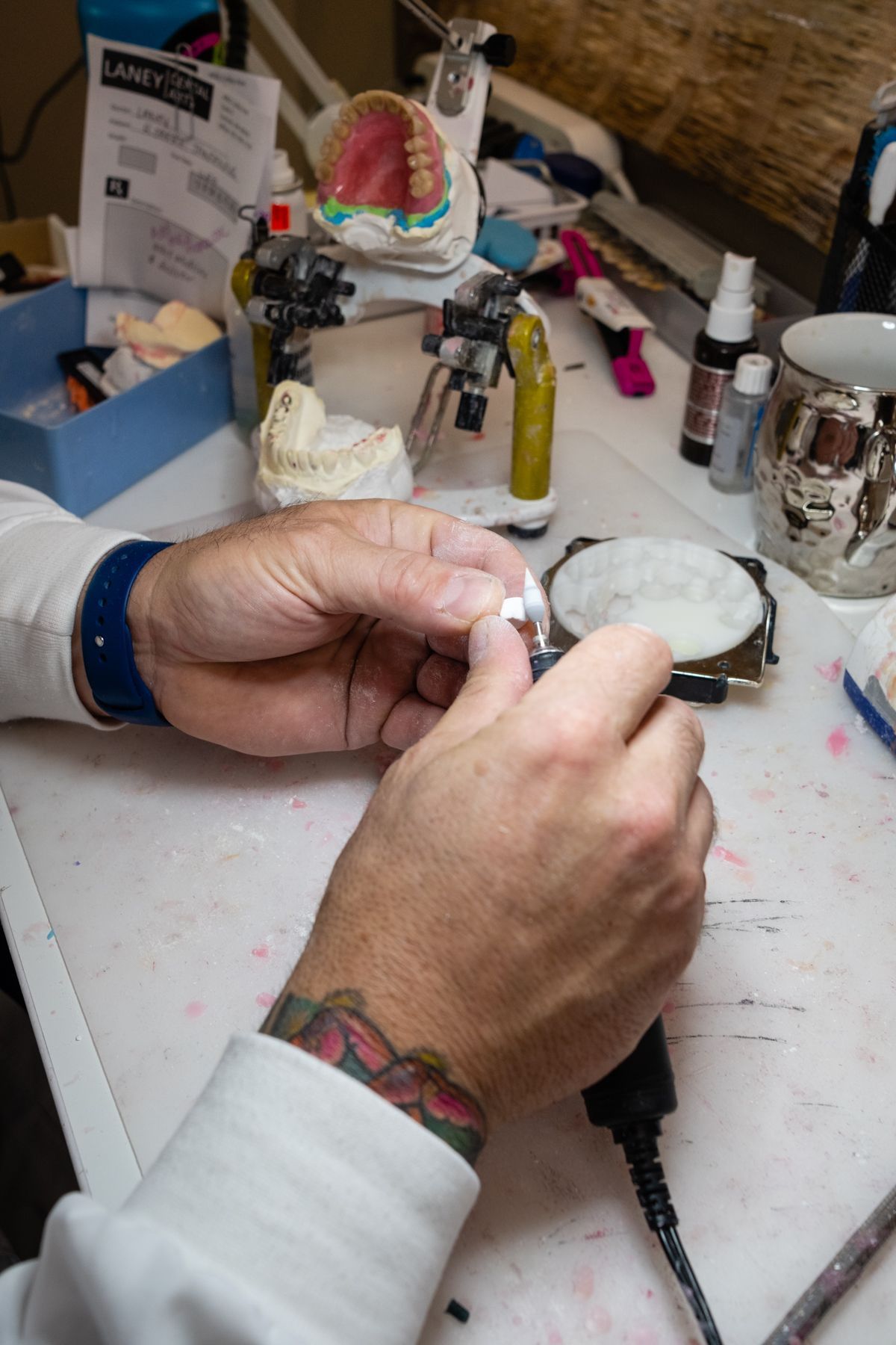 Dentist working on a tooth model, using a tool. Hands, wrist tattoo, white coat, dental lab setting.