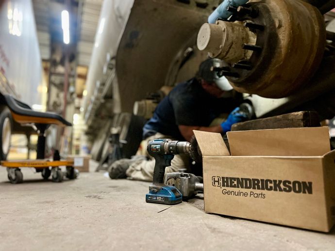 A man is working on a truck in a garage next to a box that says hendrickson genuine parts.