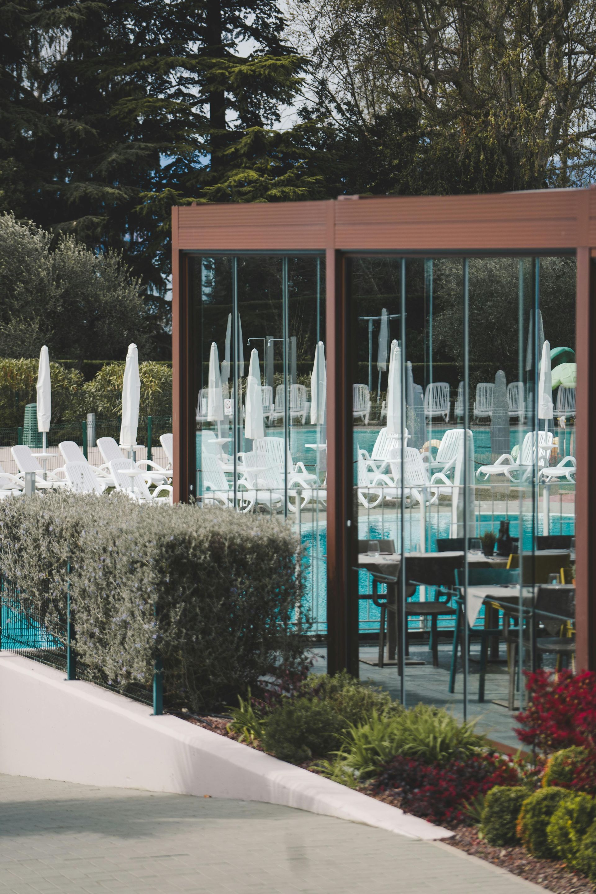 Outdoor restaurant patio with teal chairs, glass walls, closed umbrellas, and hedges by a pool