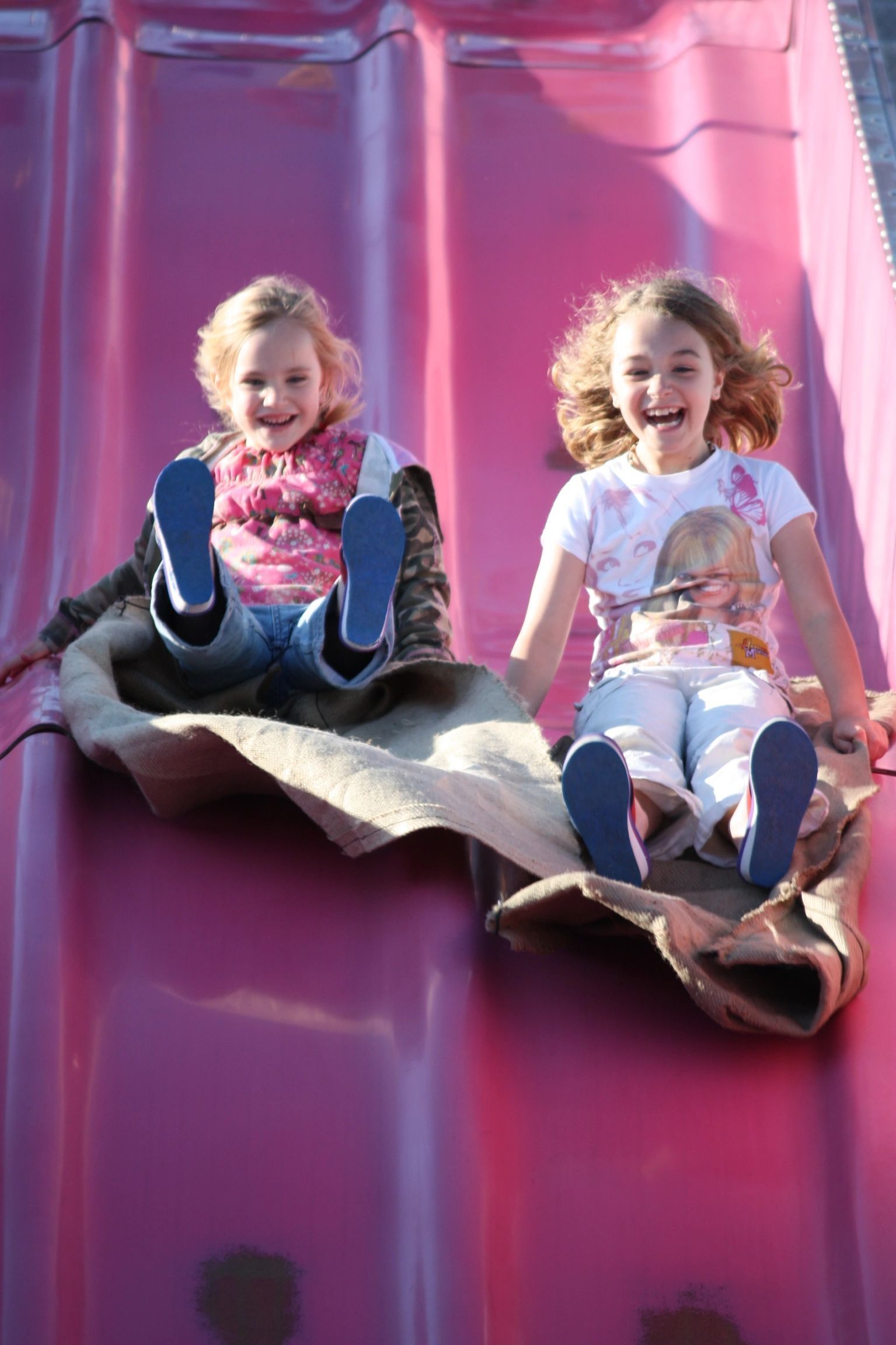 Two children sliding down a pink slide on a mat, smiling with feet out front