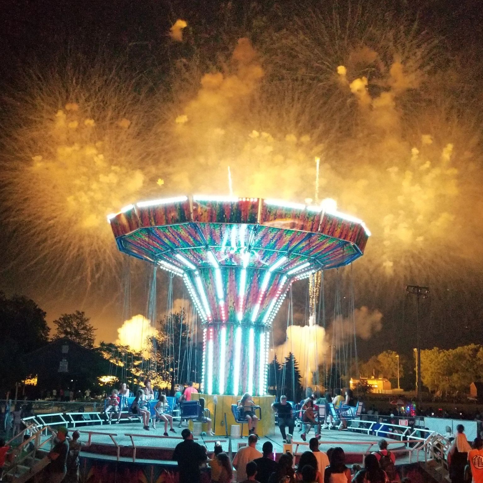Nighttime amusement ride glowing in red, blue, and white with fireworks bursting overhead