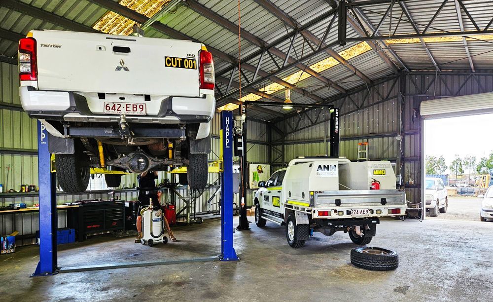 Mechanic working under a lifted ute inside a busy automotive workshop.