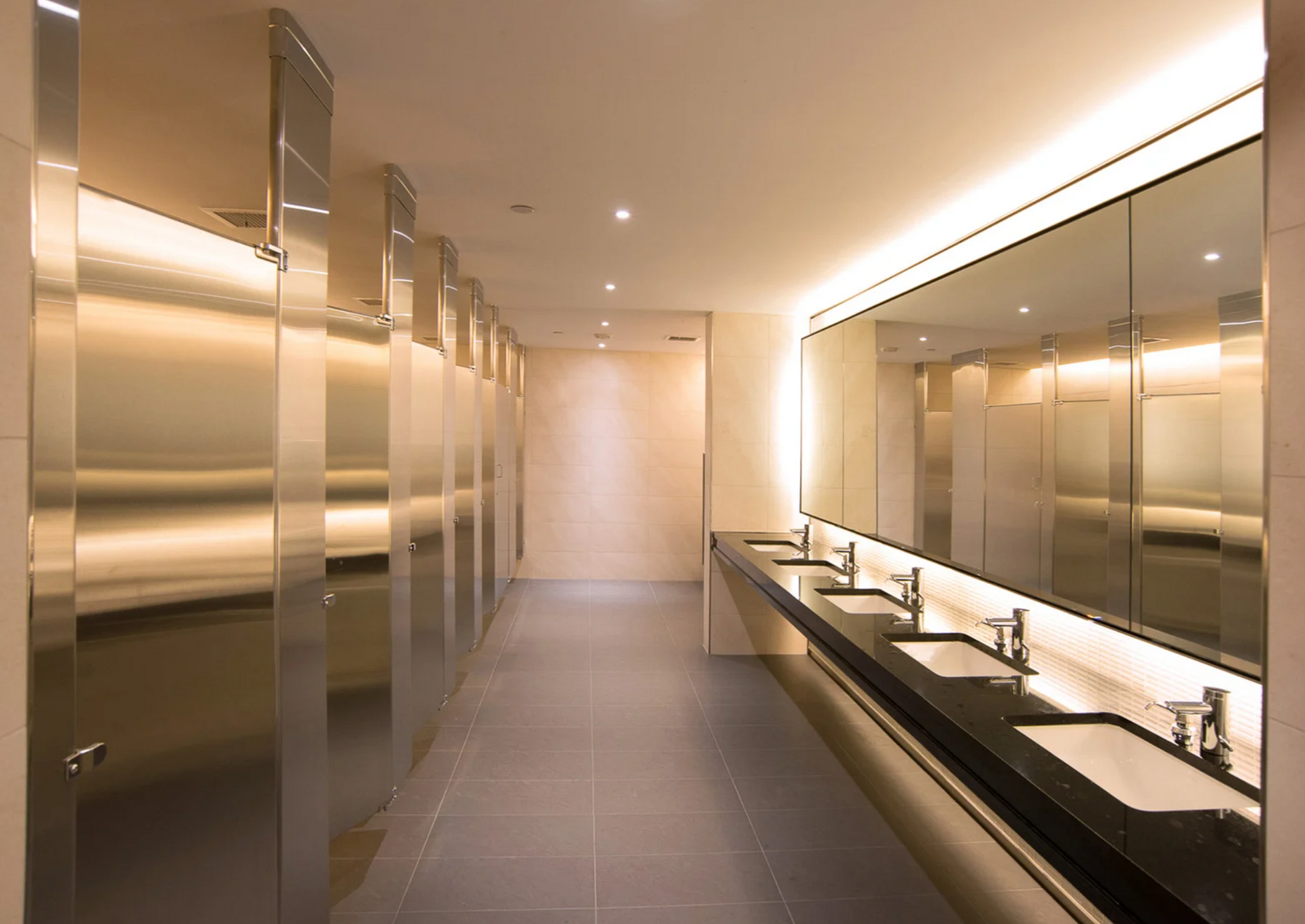 Modern public restroom with stainless steel stalls, long sink counter with mirrors, and neutral colors.