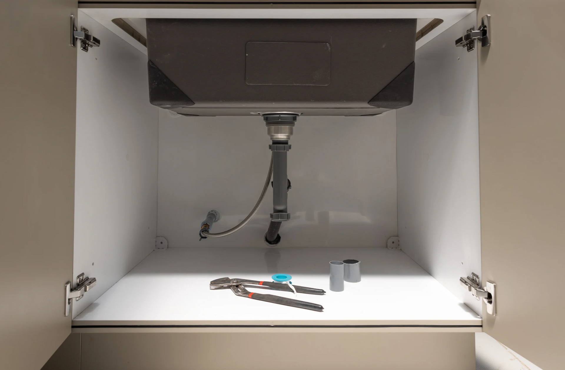 Underside of a sink in a white cabinet. Pliers, two pipe fittings, and sink drain are visible.
