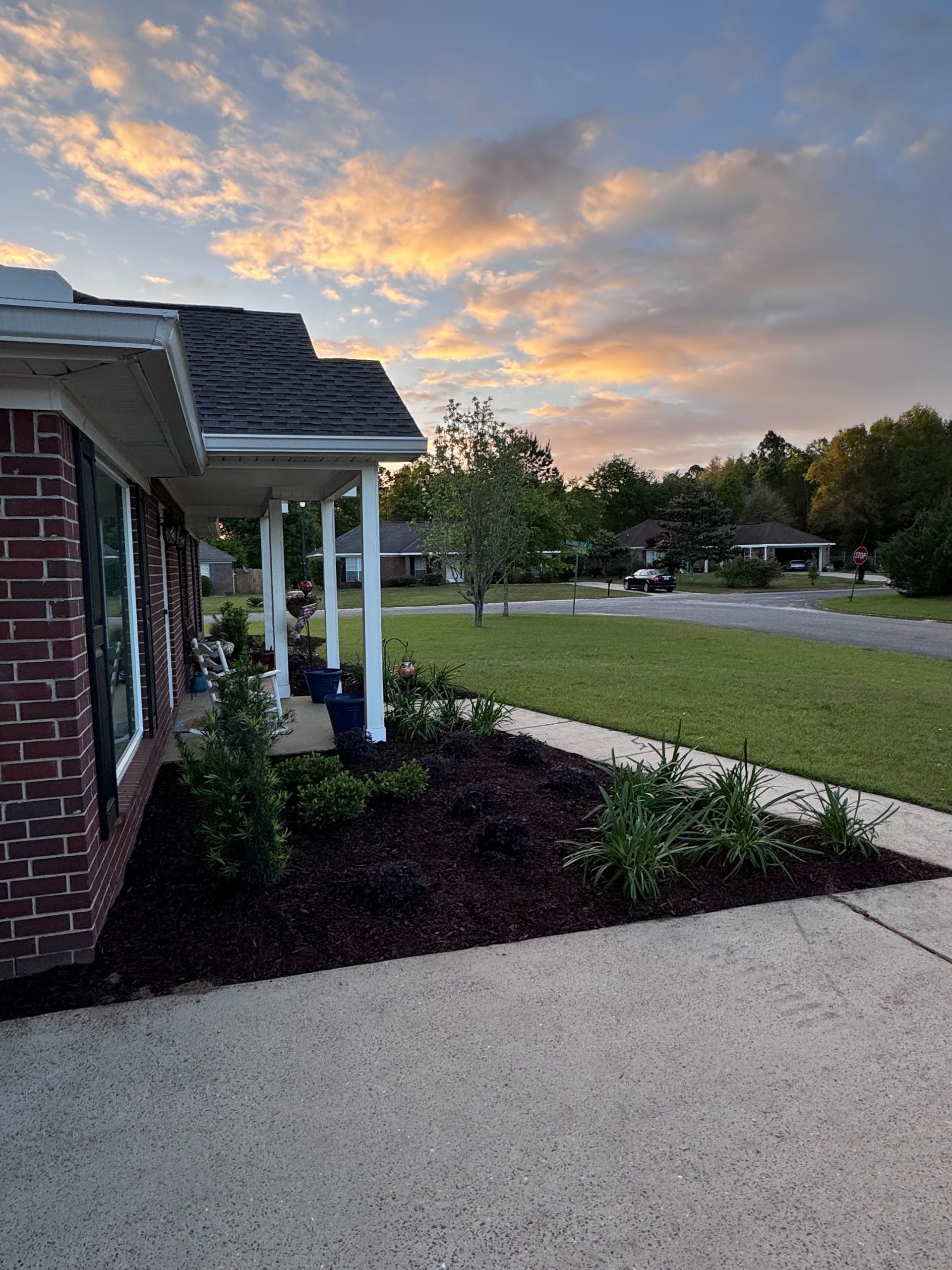 Front porch and landscaped yard at sunset, with white columns and a walkway beside the house.