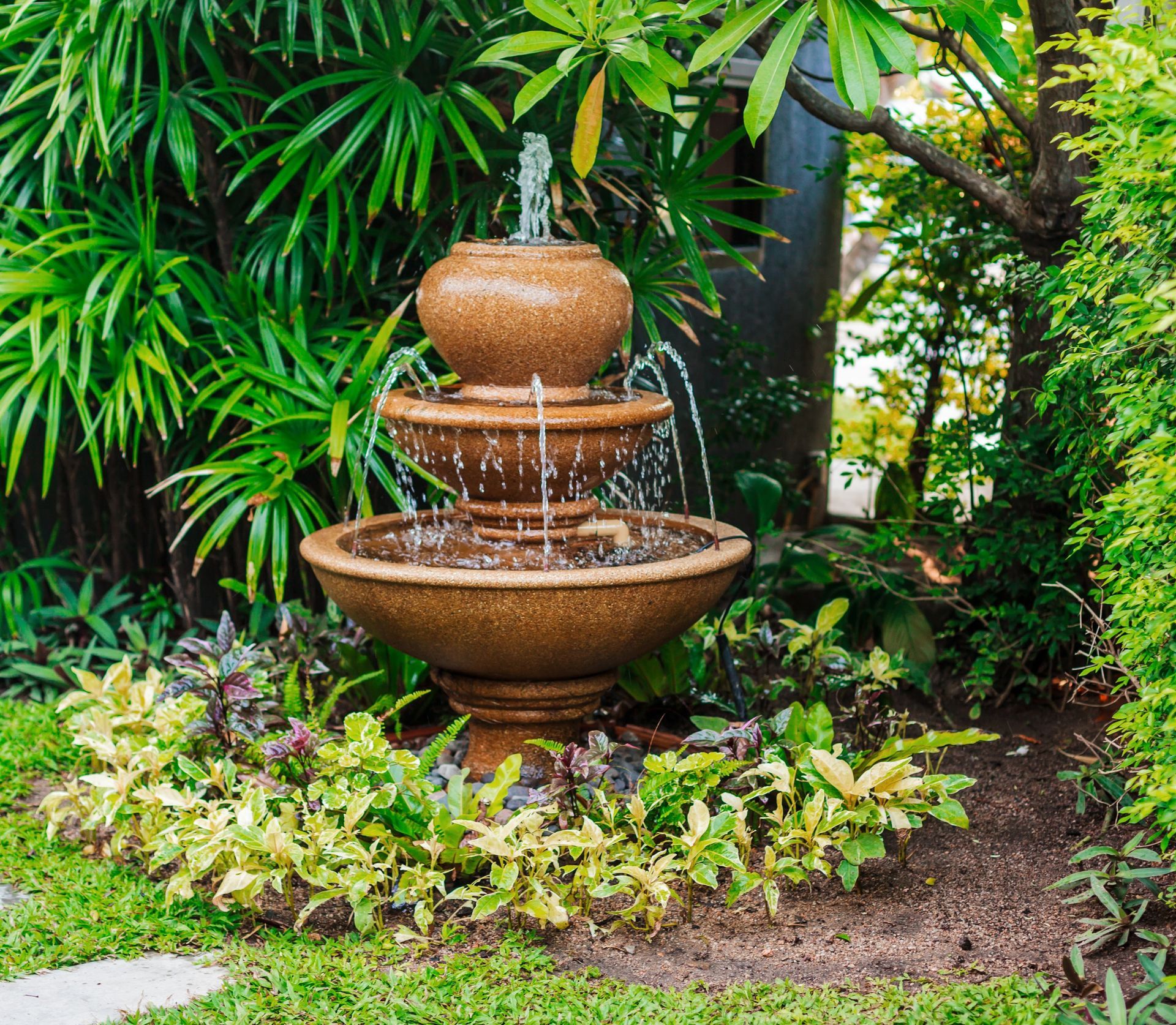 Tiered stone fountain trickling water in a lush tropical garden, surrounded by green foliage and flowers