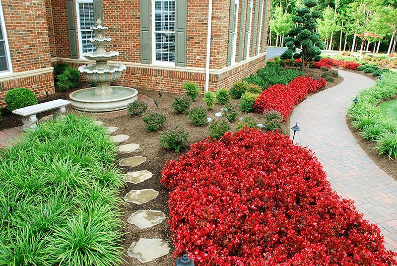 Brick house garden with red and green flower beds, stone path, and a fountain