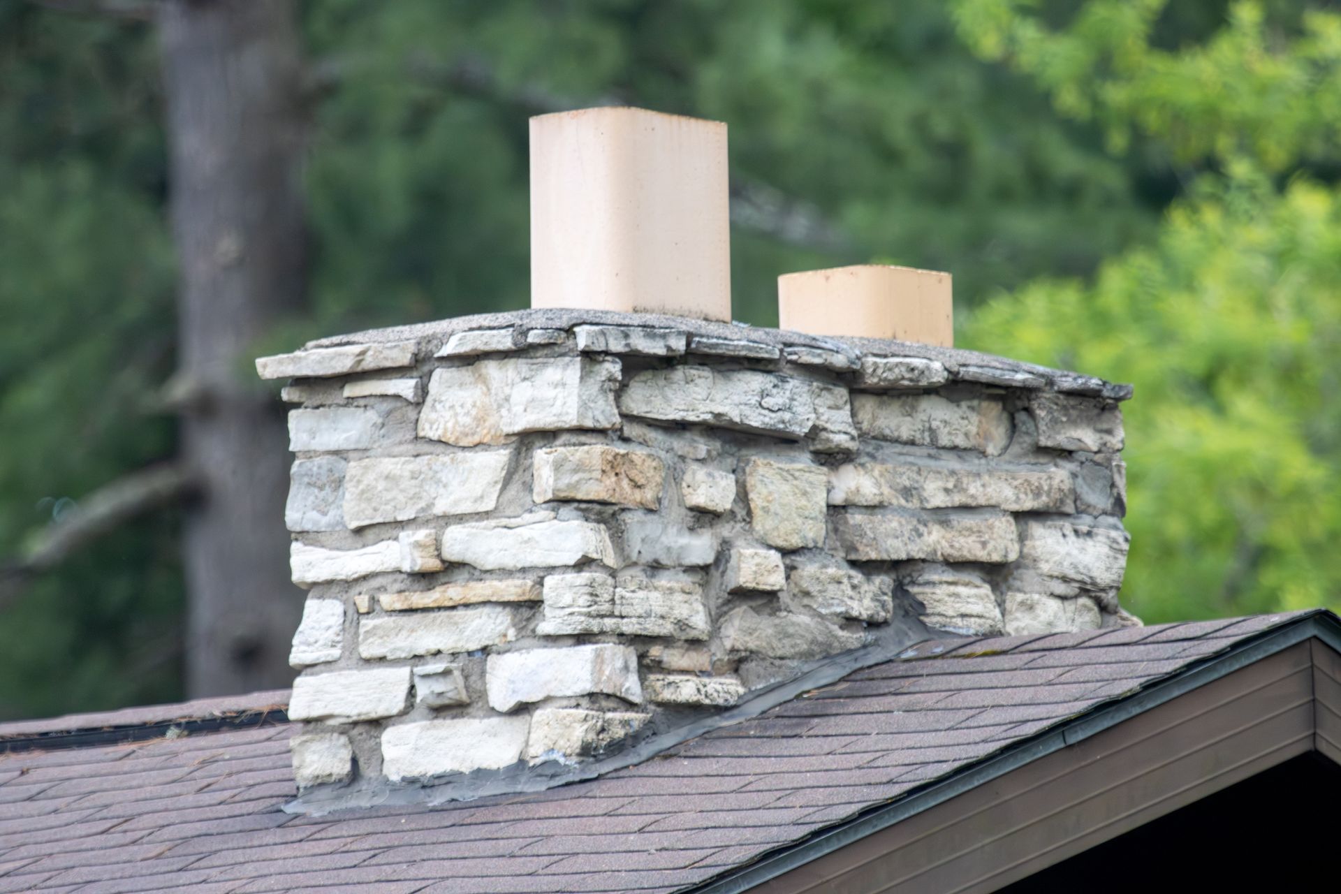 Stone chimney with two flues rising above a shingled roof and trees.