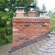 Brick chimney with three flue caps on a dark shingle roof, green trees in background.