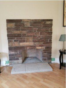 Stone fireplace in a room with hardwood floors and a small table with a lamp to the right.