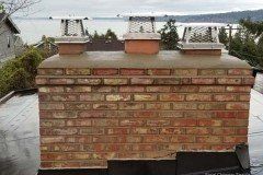 Brick chimney with three flue caps on a flat roof, overlooking a body of water and trees.