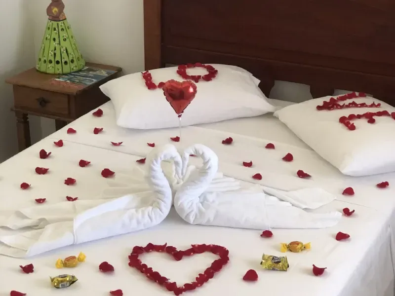 Uma cama decorada com pétalas de rosa, dois cisnes de toalha formando um coração, um balão vermelho em forma de coração e doces para um ambiente romântico.