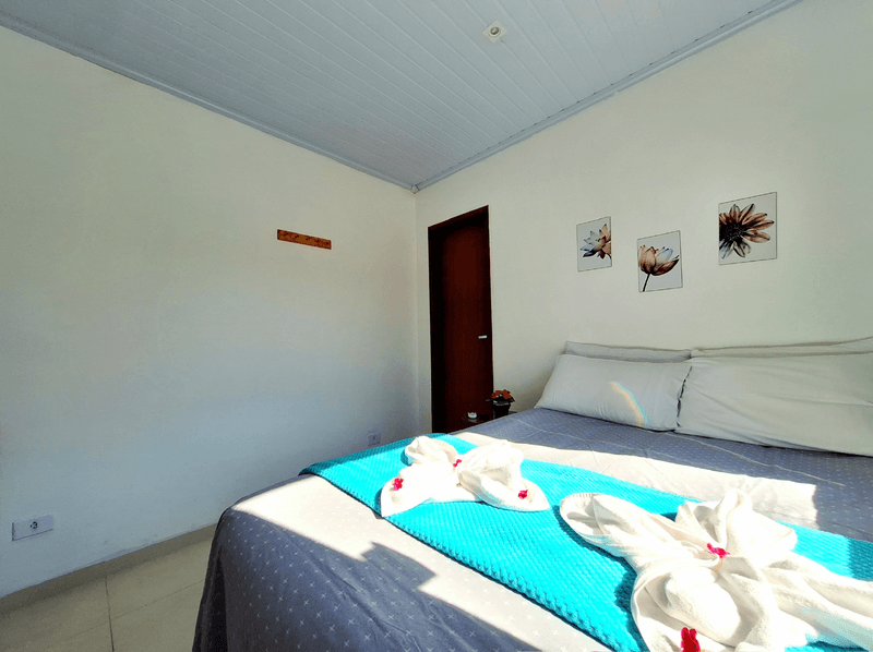 Um quarto com uma cama com travesseiros brancos e toalhas em formato de cisnes sobre um caminho de mesa azul, contra paredes brancas.