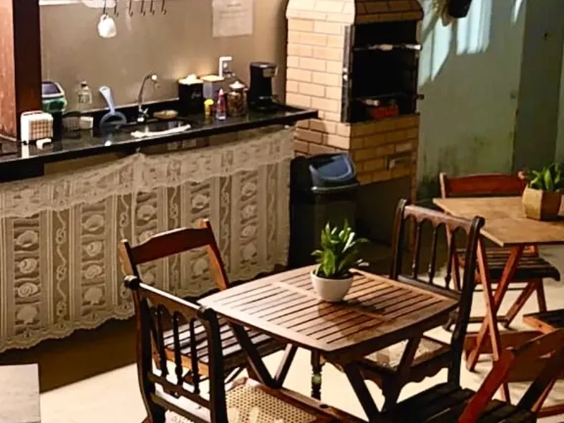 Uma aconchegante cozinha externa rústica com mesa de madeira, cadeiras, pia e churrasqueira de tijolos.