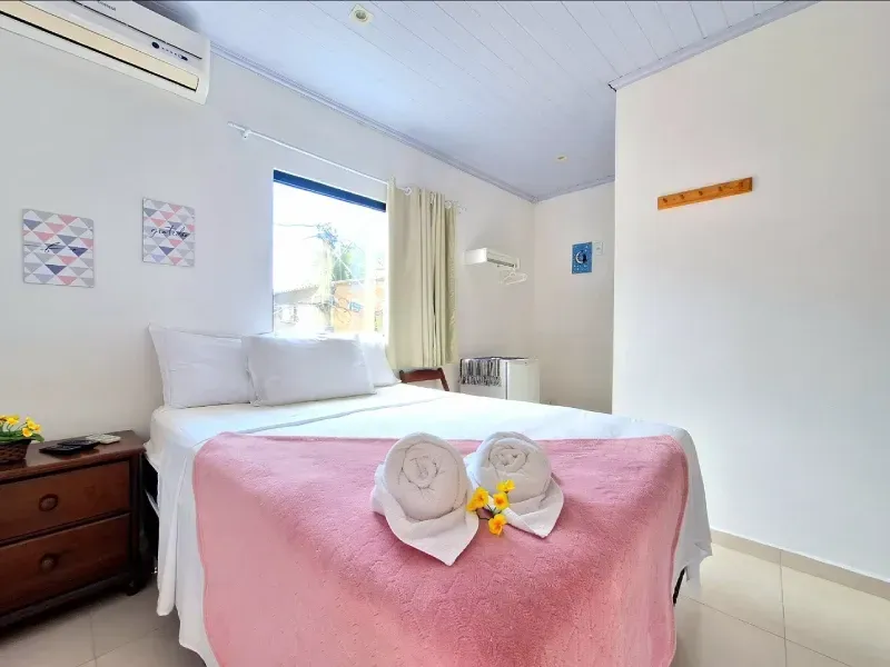 Um quarto com uma cama coberta com um cobertor rosa, travesseiros brancos, uma mesa de cabeceira e uma janela com cortinas.