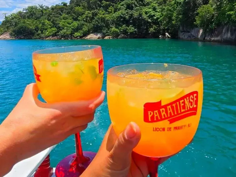 Duas pessoas brindam com coquetéis de laranja em copos personalizados, sobre as águas tropicais de um azul vibrante e uma costa verdejante.