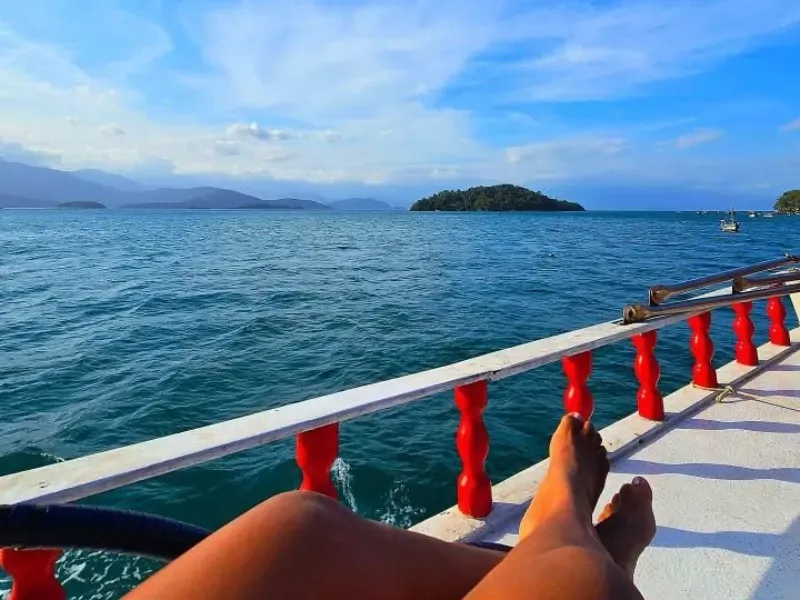 Pernas de uma pessoa apoiadas no convés branco de um barco com corrimão vermelho, com vista para um mar azul calmo e ilhas.