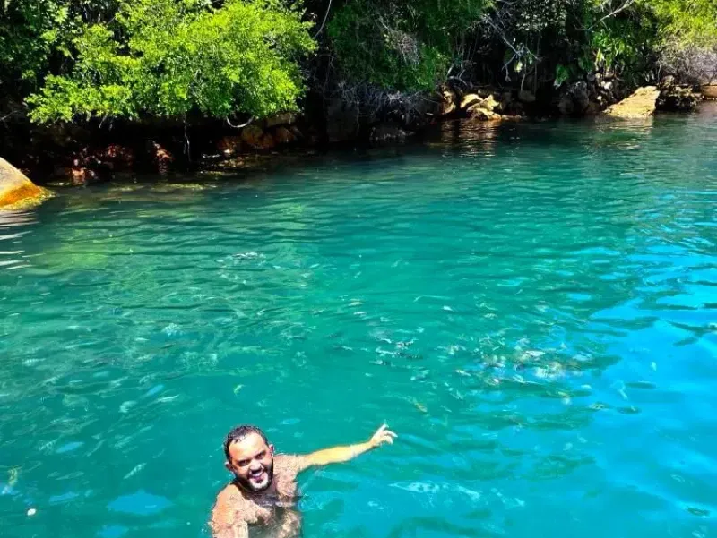 Uma pessoa sorridente nadando em águas cristalinas e vibrantes de cor turquesa perto de uma costa rochosa e exuberante.