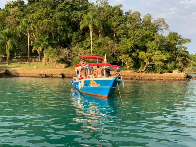 Um barco azul com toldo vermelho ancorado em águas calmas e cristalinas, com uma costa tropical exuberante e verde ao fundo.