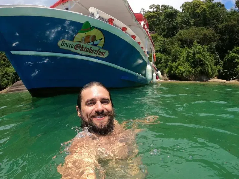 Uma pessoa barbuda sorri na água verde e cristalina em frente a um barco azul chamado