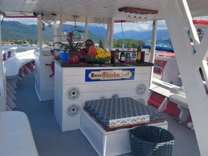Um convés de barco branco com um pequeno bar de madeira com bebidas, frutas e alto-falantes, sob um toldo com vista para o oceano.