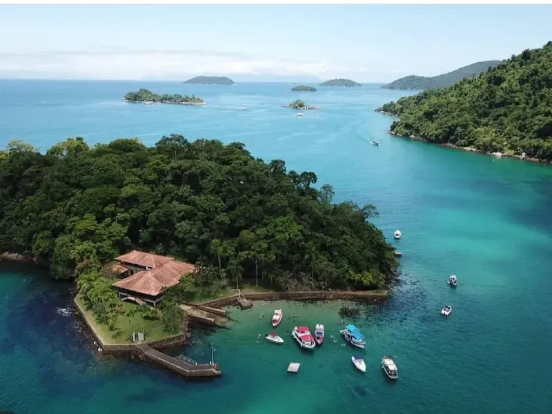 Vista aérea de uma ilha arborizada com uma casa de telhado vermelho e vários barcos pequenos ancorados em águas azuis cristalinas.