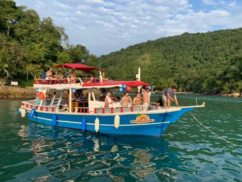 Um barco de festa azul brilhante com toldo vermelho flutua em águas calmas e verdes perto de uma costa exuberante e arborizada.