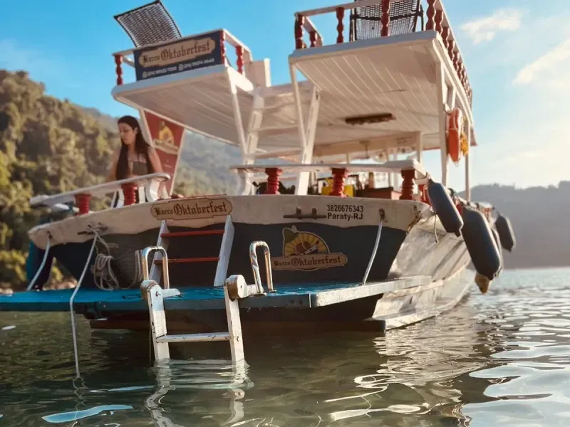 Um barco atracado em águas calmas perto de uma costa montanhosa e arborizada, com uma pessoa em pé no convés traseiro.