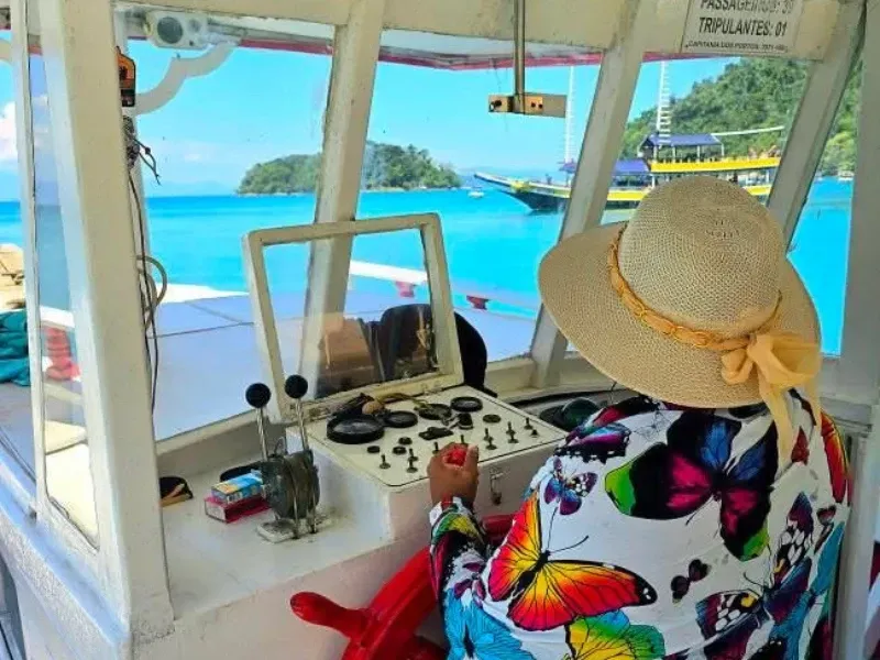 Uma pessoa usando um chapéu de sol e uma blusa com estampa de borboletas conduz um barco em direção a uma ilha tropical em águas cristalinas.