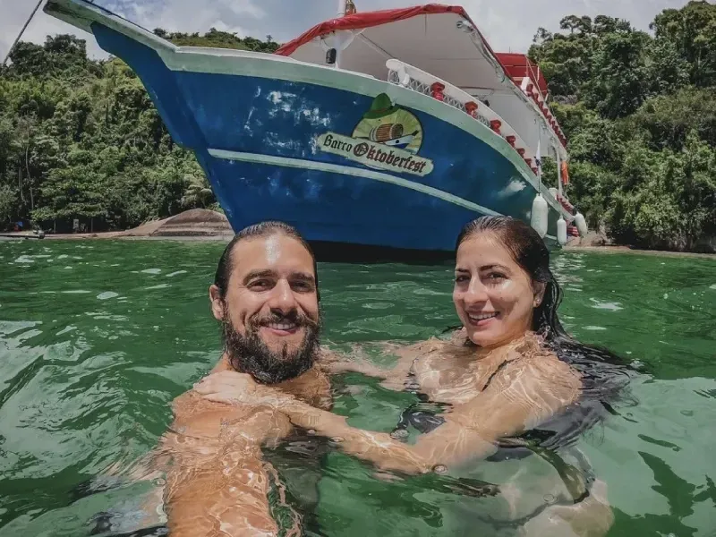 Um casal sorridente posa para uma selfie enquanto nada em águas cristalinas e verdes, com um barco azul e branco ao fundo.