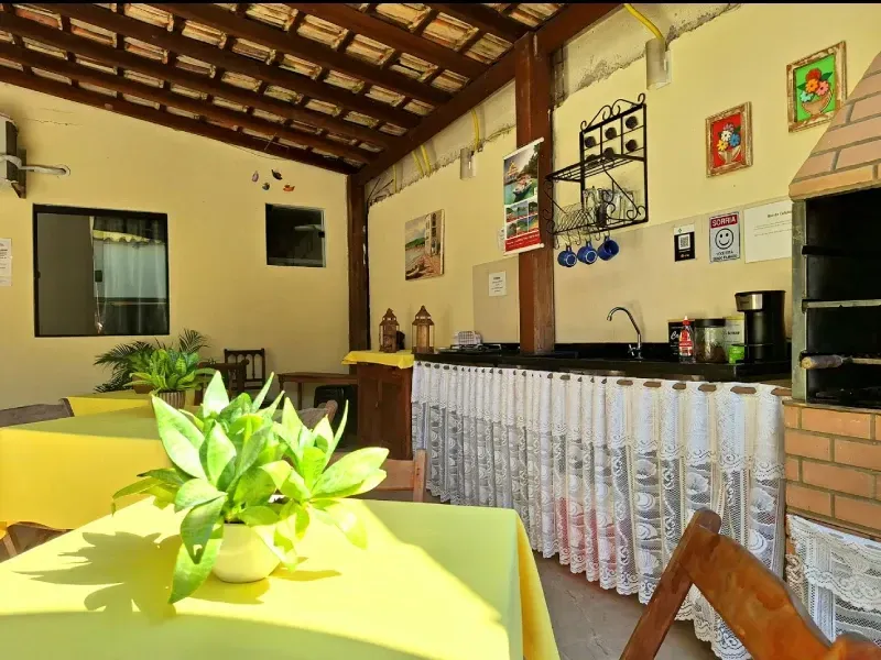 Uma área de jantar rústica no pátio, com mesas cobertas com toalhas amarelas, vasos de plantas, um balcão de cozinha com cortinas e uma churrasqueira.