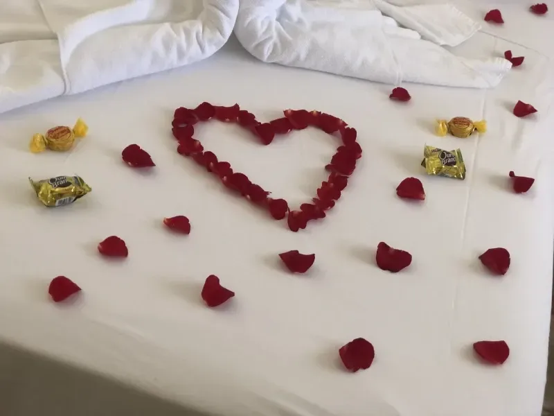 Uma cama de hotel branca decorada com um coração feito de pétalas de rosas vermelhas, pétalas espalhadas e quatro chocolates embalados individualmente.